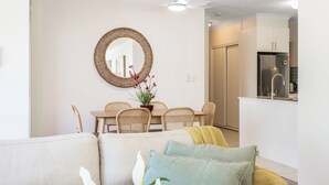 Een moderne woonkamer met een bank, kussens, een eettafel met stoelen, een ronde spiegel en een keuken met een koelkast.