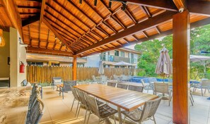 Un patio exterior con mesas y sillas de madera, una piscina y una zona cubierta con techo de madera.