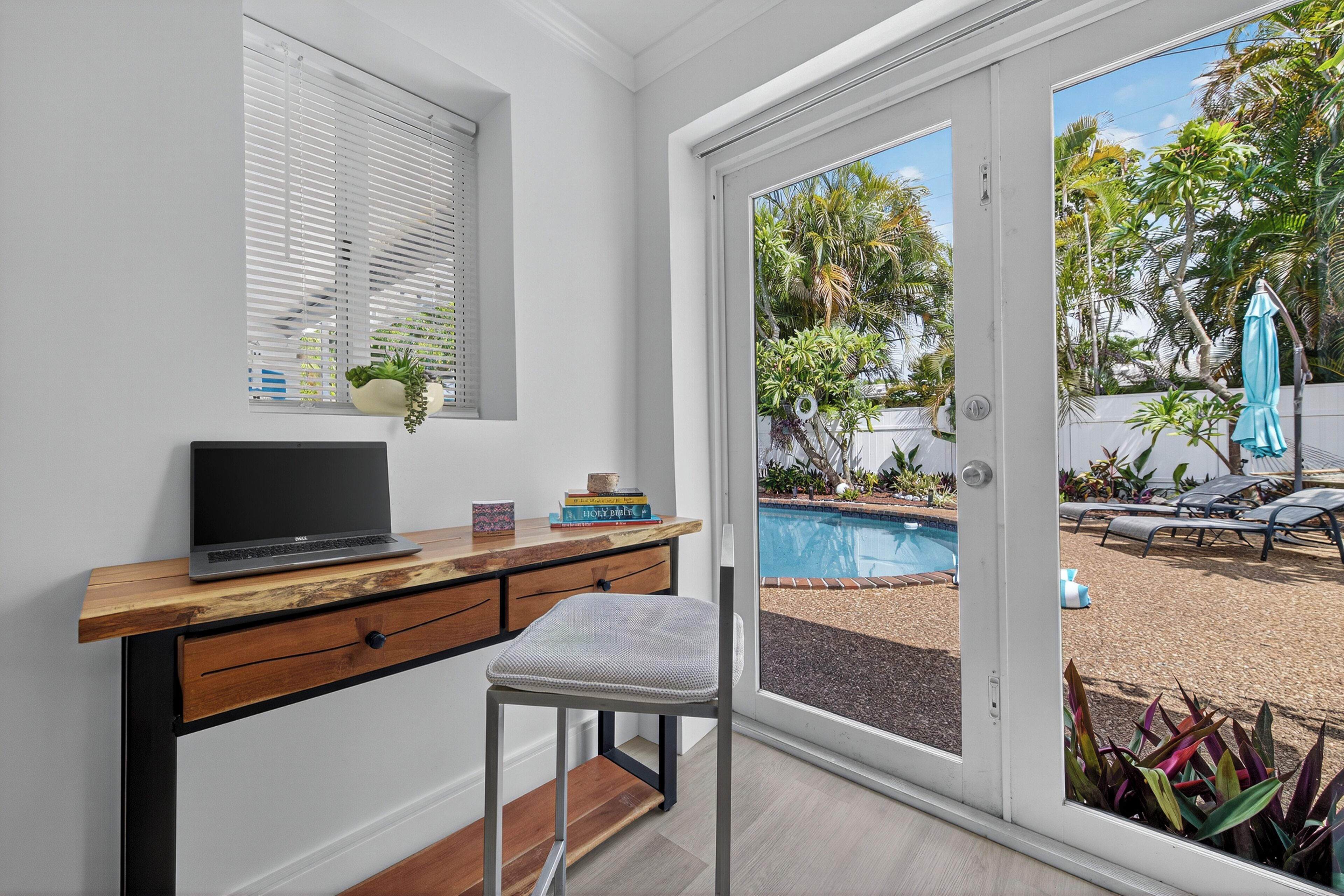 Ein Zimmer mit einem Holztisch, einem Laptop, einem Stuhl, einem Fenster mit Jalousien und Blick auf einen Pool und Gartenmöbel.