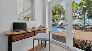 A room with a wooden desk, a laptop, a chair, a window with blinds, and a view of a pool and outdoor furniture.