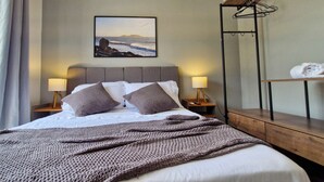 Un lit bien fait, avec une couverture tricotée, deux oreillers et un tableau représentant une scène de plage accroché au mur.