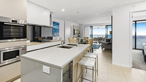 A modern kitchen with built-in appliances, a central island with bar stools, and a dining area with a view. - Lovely 2 Bedroom Holiday Ocean View Unit — Q Stay (Surfers Paradise)