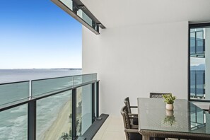 Un balcon avec une table et des chaises en verre, donnant sur une plage et l’océan.