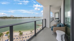 A balcony with a view of a beach and ocean. There are lounge chairs and a small table.