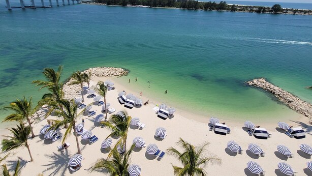 A beach with clear turquoise water, a bridge in the background, and multiple umbrella-covered seating areas.