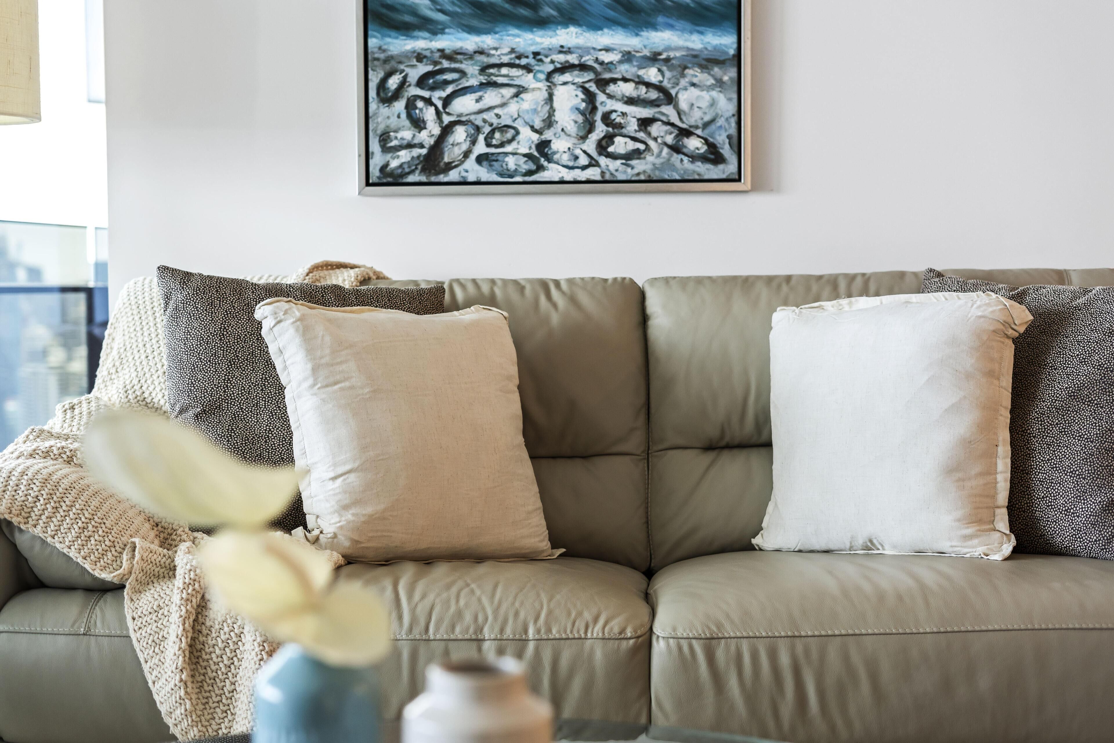 Een beige leren bank met twee kussens, een vaas met bloemen en een kunstwerk in een lijst aan de muur.