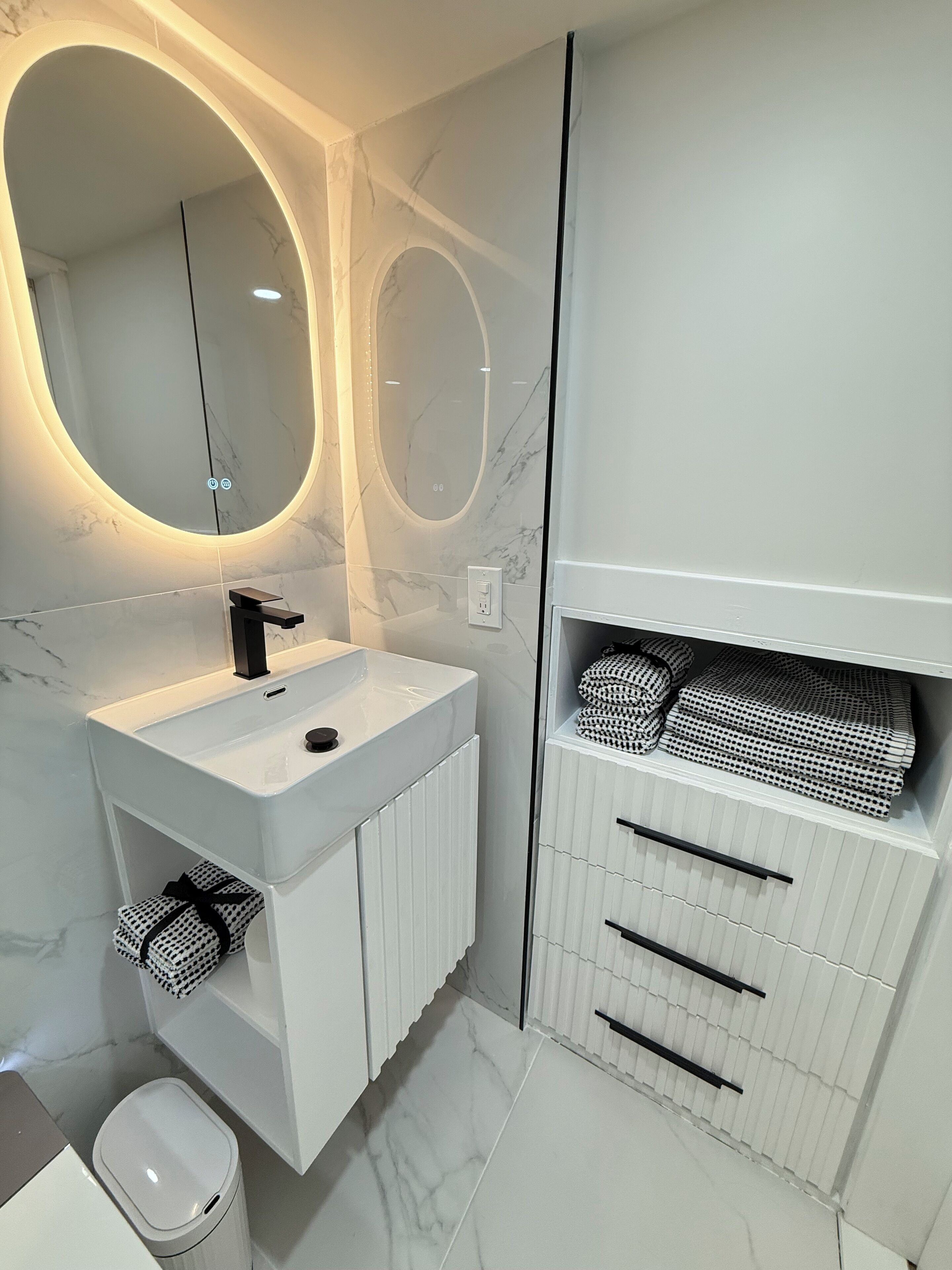 Une salle de bain moderne avec un meuble-lavabo blanc, un miroir rond et une étagère intégrée où sont rangées des serviettes à motifs.