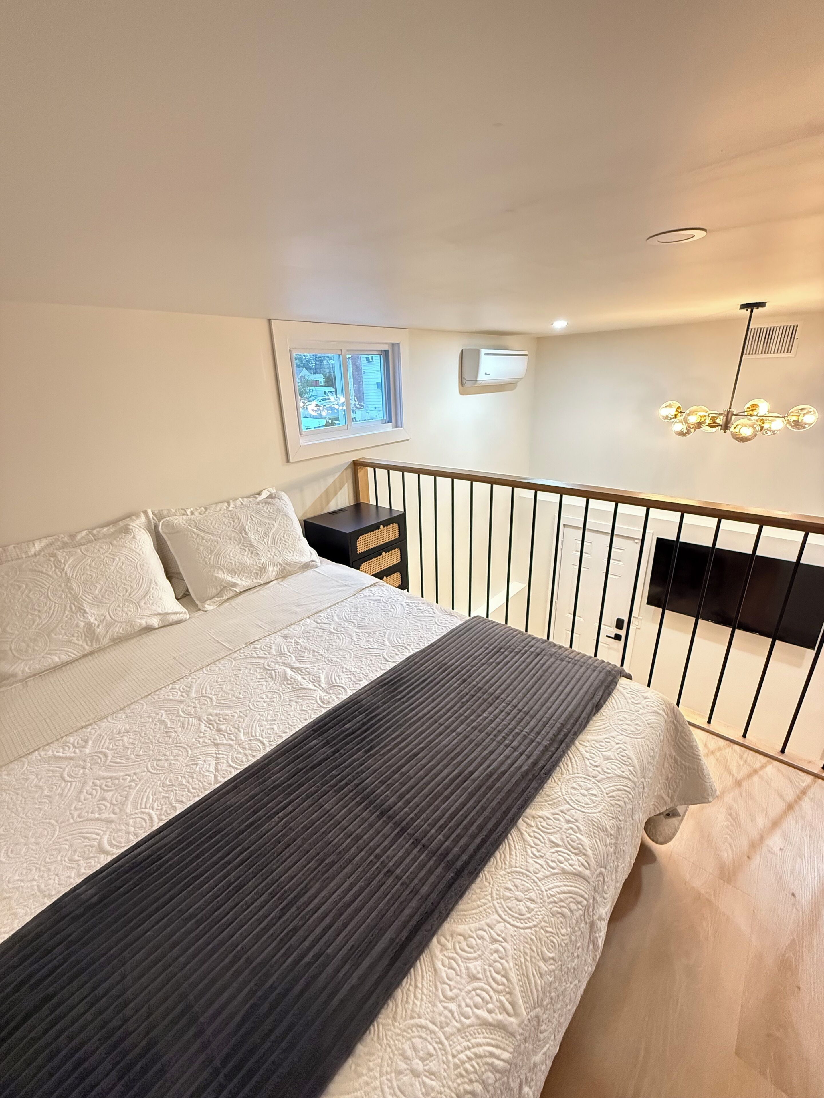 A bedroom with a bed, a window, an air conditioner, a ceiling light, and a railing.