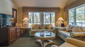 A hotel room with a sofa set, a television, a coffee table, and large windows offering a view of trees.