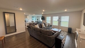 Ein geräumiges Wohnzimmer mit einem großen grauen Ecksofa, einem Couchtisch und einer kleinen Holzbank. Im Hintergrund ist eine Küche mit Esstisch und Stühlen zu sehen.
