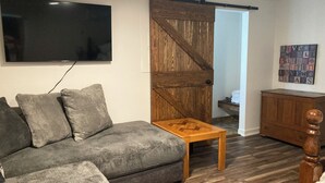 Een woonkamer met een grijze bank, een houten salontafel, een aan de muur bevestigde televisie, een houten deur en een kast.