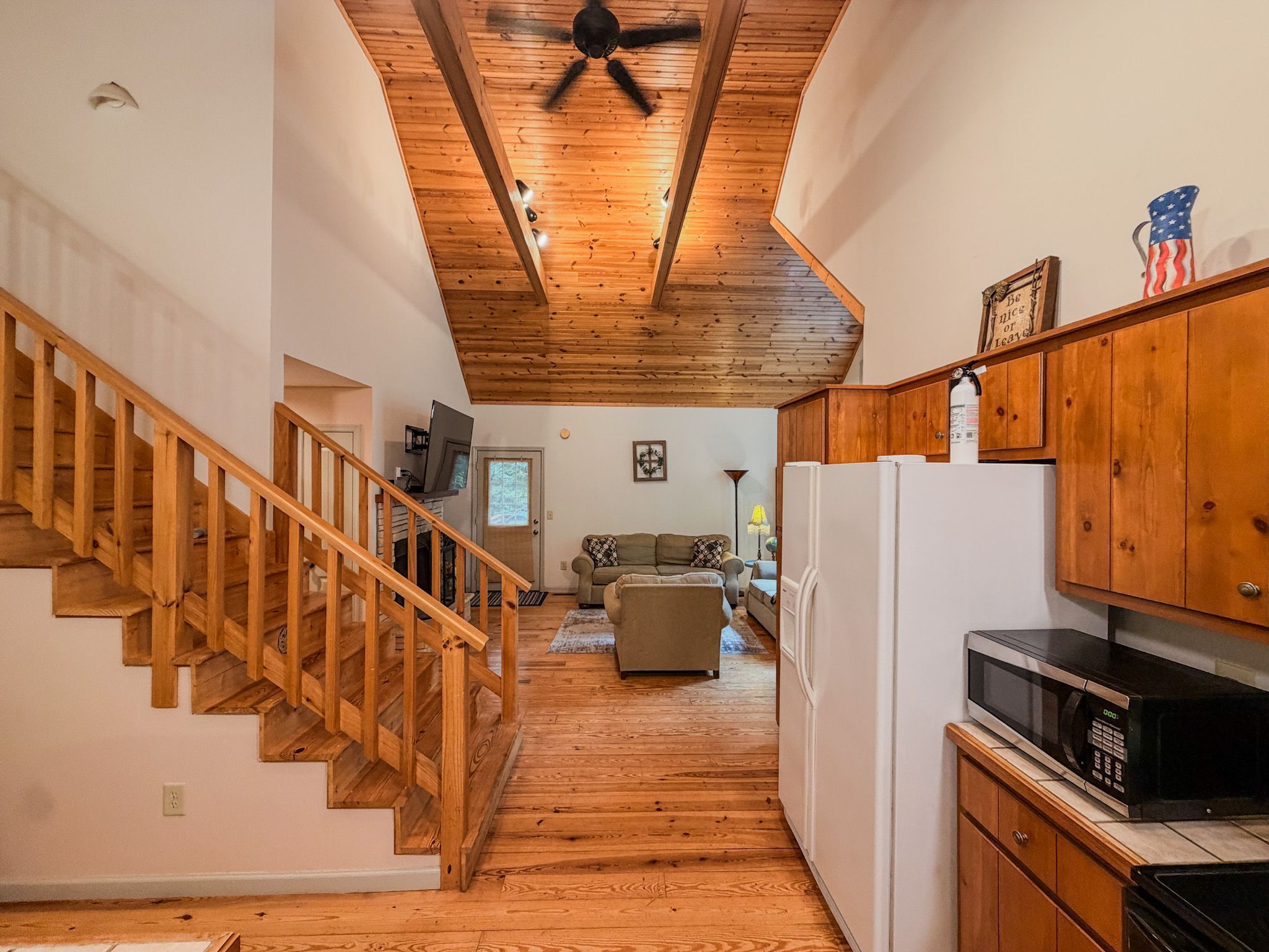 Una escalera de madera conduce a una sala de estar con ventilador de techo, una cocina con microondas y refrigerador.