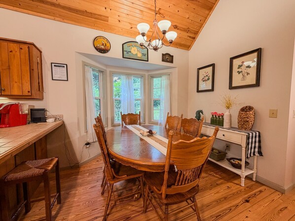A dining area with a wooden table, chairs, and a chandelier. There are windows with curtains, a kitchen area with a coffee maker, and a framed picture on the wall.