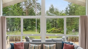 A conservatory with a grey sofa and cushions, large windows offering a view of a lush garden. - Beautiful home in Ebeltoft with WiFi (Ebeltoft)