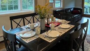 A dining area with a black table set for four, surrounded by black chairs. There is a vase with flowers on the table, and a black leather couch in the background.