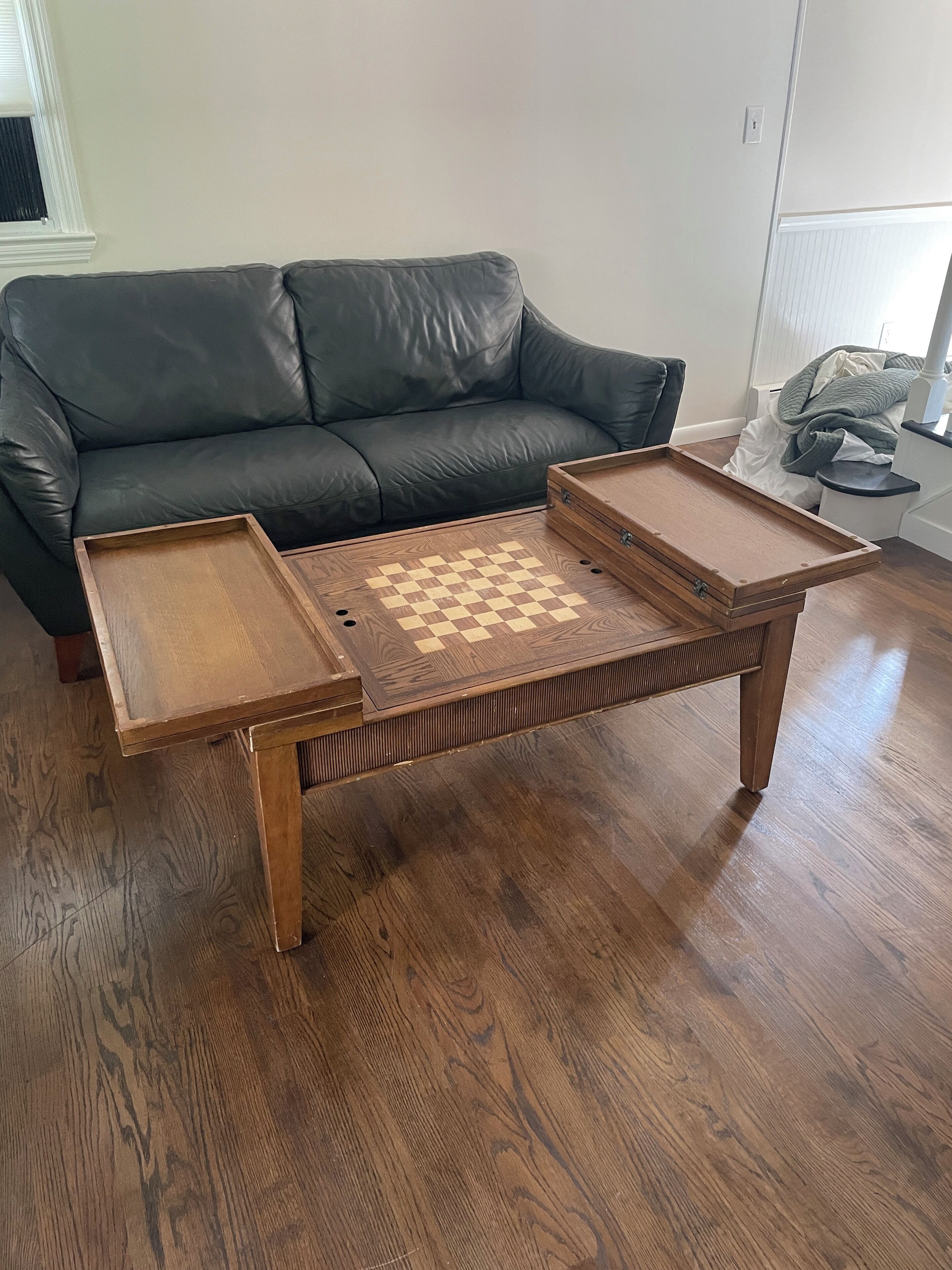 Una mesa de centro de madera con un tablero de ajedrez en el centro, ubicada sobre un piso de parqué en un salón con un sofá de cuero verde oscuro.