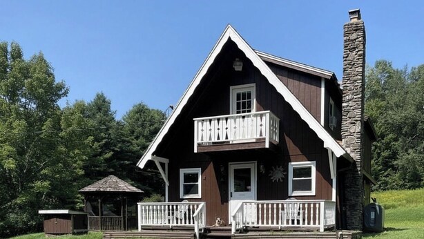 A two-story wooden house with a stone chimney, a balcony, and a small porch.
