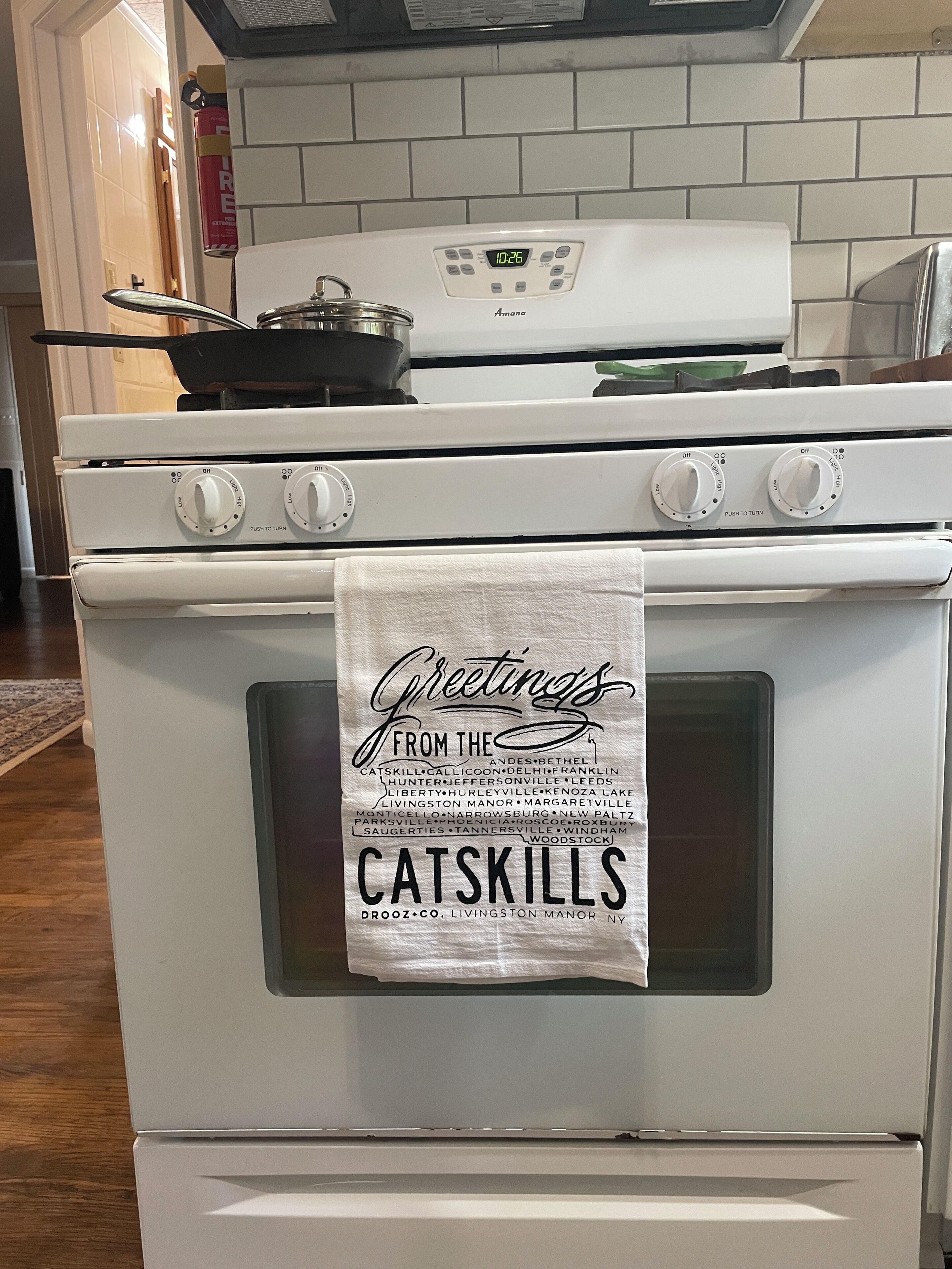 Una cocina con estufa blanca, un reloj digital y una toalla colgada en la puerta del horno con la inscripción 'Greetings from the Catskills'.