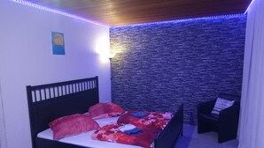 A bedroom with a bed, a chair, a wall with a stone pattern, and a ceiling with wooden panels and LED lights.