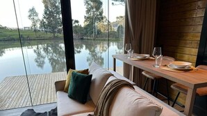 Une pièce moderne avec une table en bois dressée pour le repas, un canapé avec des coussins et une grande fenêtre donnant sur une étendue d’eau.