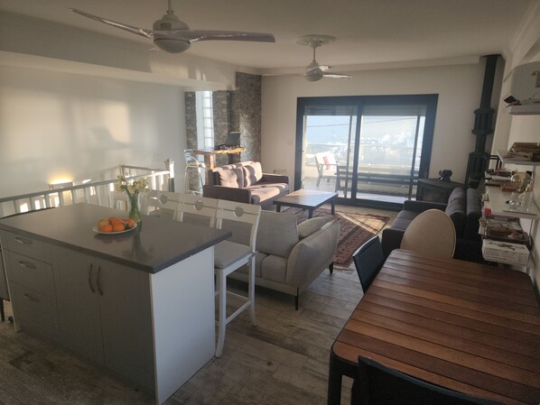 A modern kitchen with a central island, dining area with a wooden table, and a living space with a sofa and a fireplace.