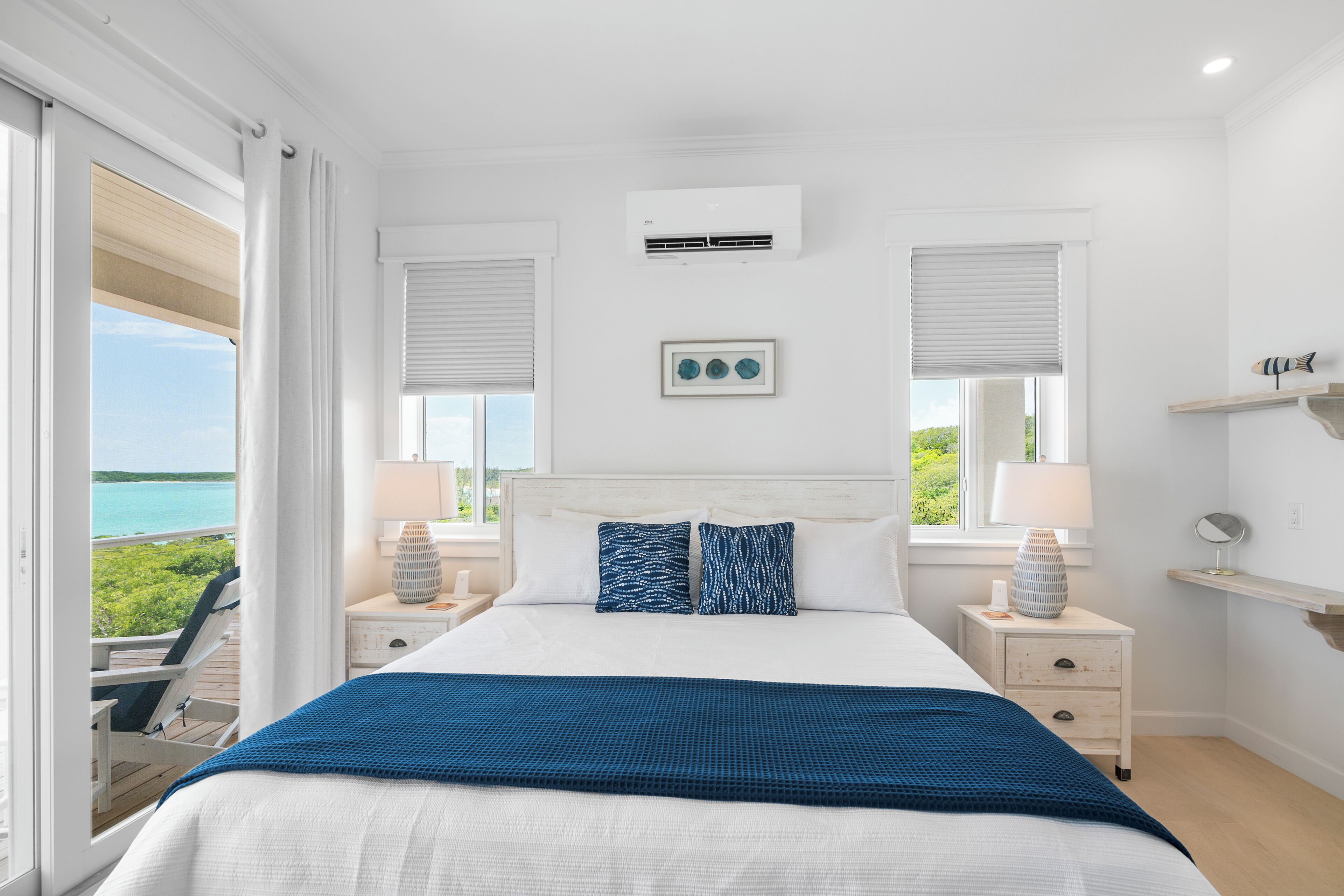 A bedroom with a bed, two bedside tables, a nightstand, a window with a view of the sea, and an air conditioning unit.