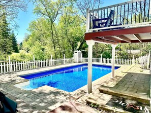 Une piscine rectangulaire entourée d’une clôture blanche, située dans un espace aménagé avec des arbres et une terrasse couverte.