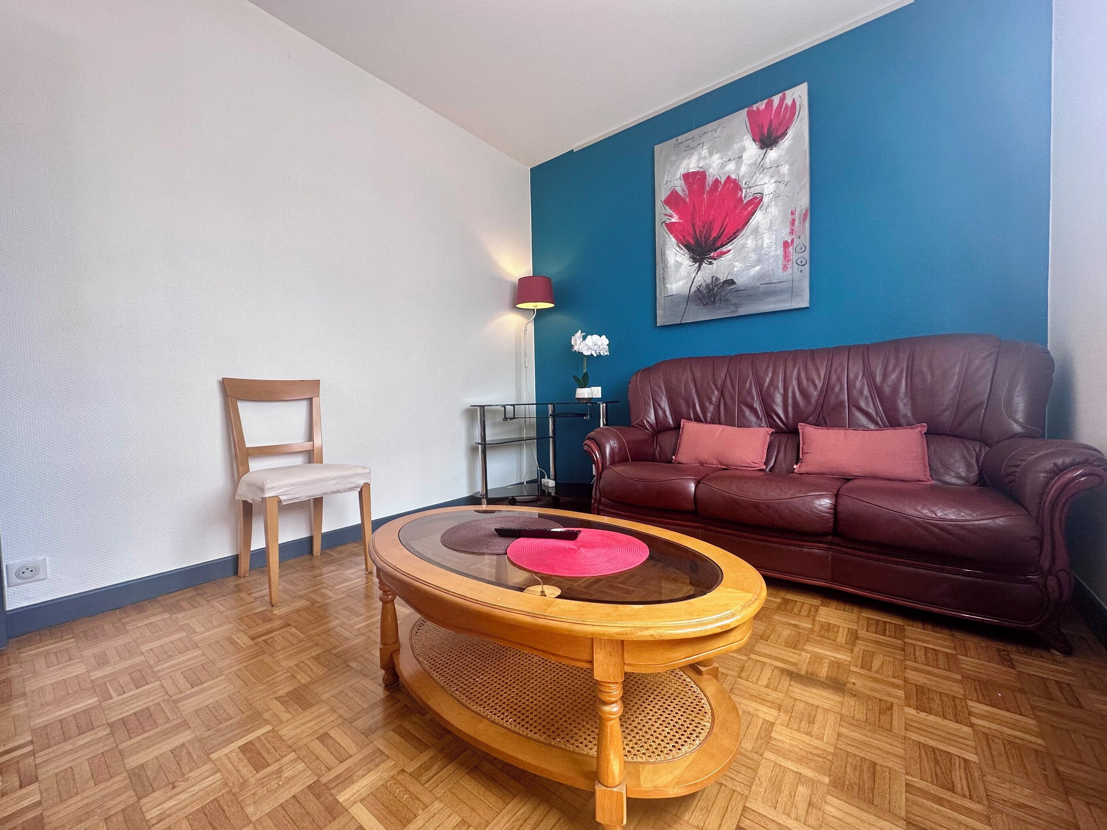 A living room with a brown leather sofa, a wooden chair, a round wooden coffee table, and a blue accent wall with a floral painting.