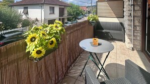 Un balcon avec une table et des chaises, un pot de fleurs contenant des tournesols, et une vue sur une maison et une rue en arrière-plan.