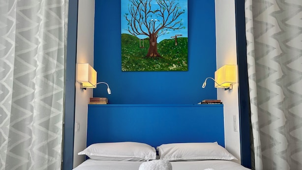 Une chambre d’hôtel avec un lit, des oreillers blancs, une tête de lit bleue, un tableau représentant un arbre et deux lampes fixées au mur.