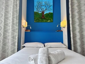 Hotelkamer met een bed, witte lakens, twee kussens, een blauw hoofdeinde, een schilderij van een boom, twee wandlampen en een raam met gordijnen met een patroon.