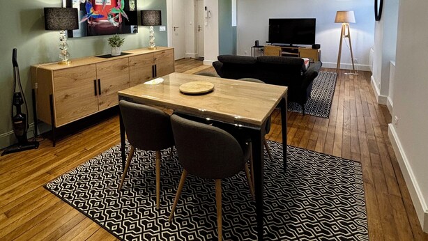 Una sala de estar moderna con una mesa de comedor de madera, dos sillas, un sofá, un televisor y un tapete estampado.