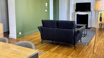 Een woonkamer met een donkere bank, een open haard, een televisie en een houten vloer.