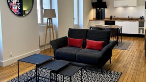 A modern living room with a sofa, a small table, a floor lamp, and a kitchen area with a stove and cabinets.