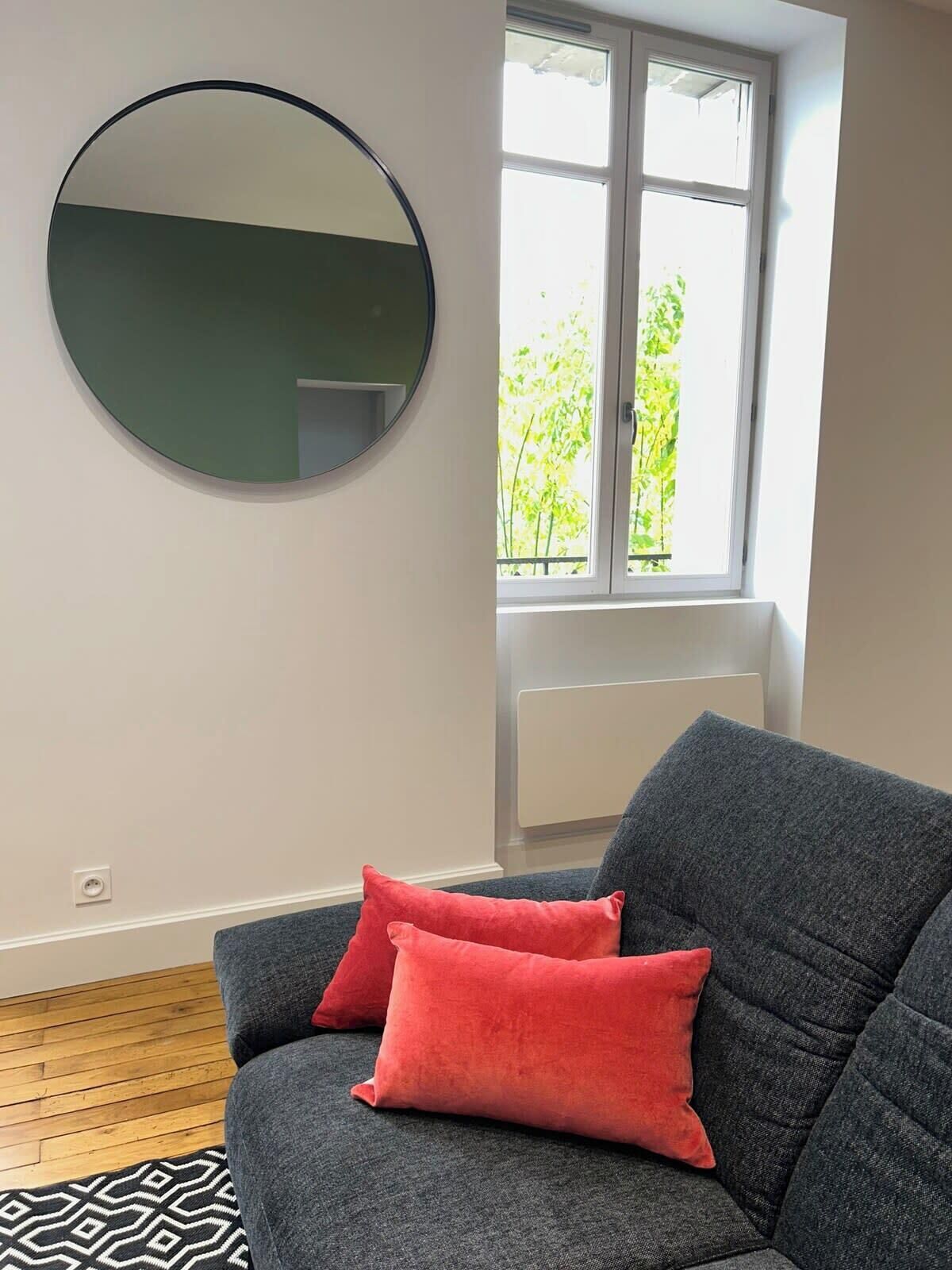 Un salon moderne avec un canapé gris, deux coussins rouges, un miroir rond et une fenêtre donnant sur un paysage verdoyant.
