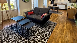Ein modernes Wohnzimmer mit einer Couch, einem Couchtisch und einem gemusterten Teppich. Im Hintergrund ist eine Küche mit weißen Schränken und ein Essbereich mit Stühlen zu sehen.