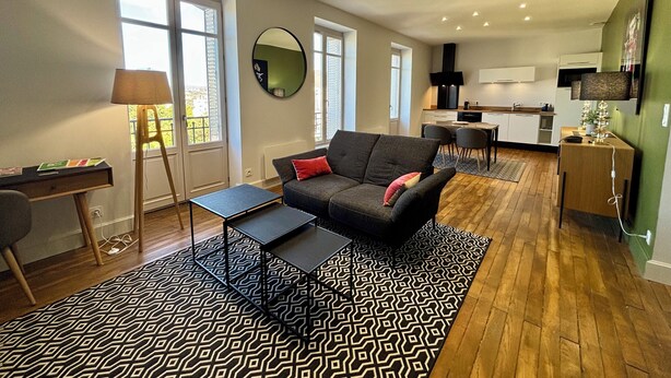 Ein modernes Wohnzimmer mit einer Couch, einem Couchtisch und einem gemusterten Teppich. Es gibt einen Essbereich mit Stühlen und eine Küche im Hintergrund.