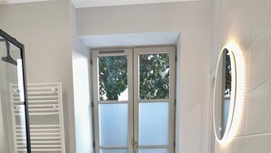 Ein Badezimmer mit Duschkabine aus Glas, einem Fenster mit satiniertem Glas, einem schwarzen Stuhl und einem Stapel gelber Handtücher auf einer weißen Ablagefläche.