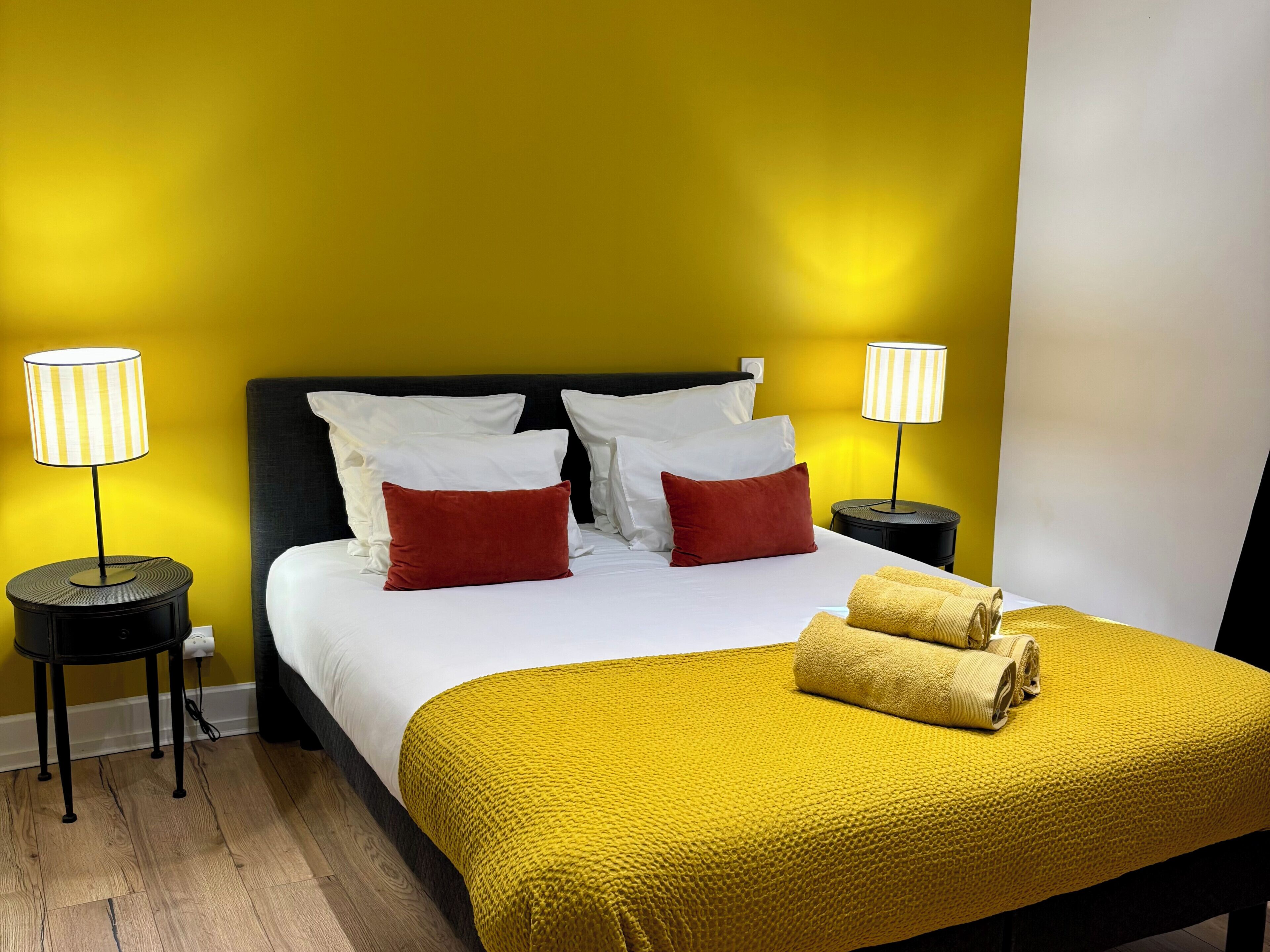 Une chambre d’hôtel avec un lit, deux lampes de chevet, une couverture jaune et un oreiller rouge.