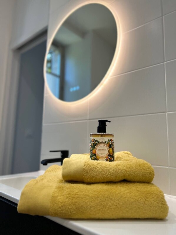 Une salle de bain avec un lavabo blanc, une serviette jaune et une bouteille d’huile essentielle Panier des Sens Provence.