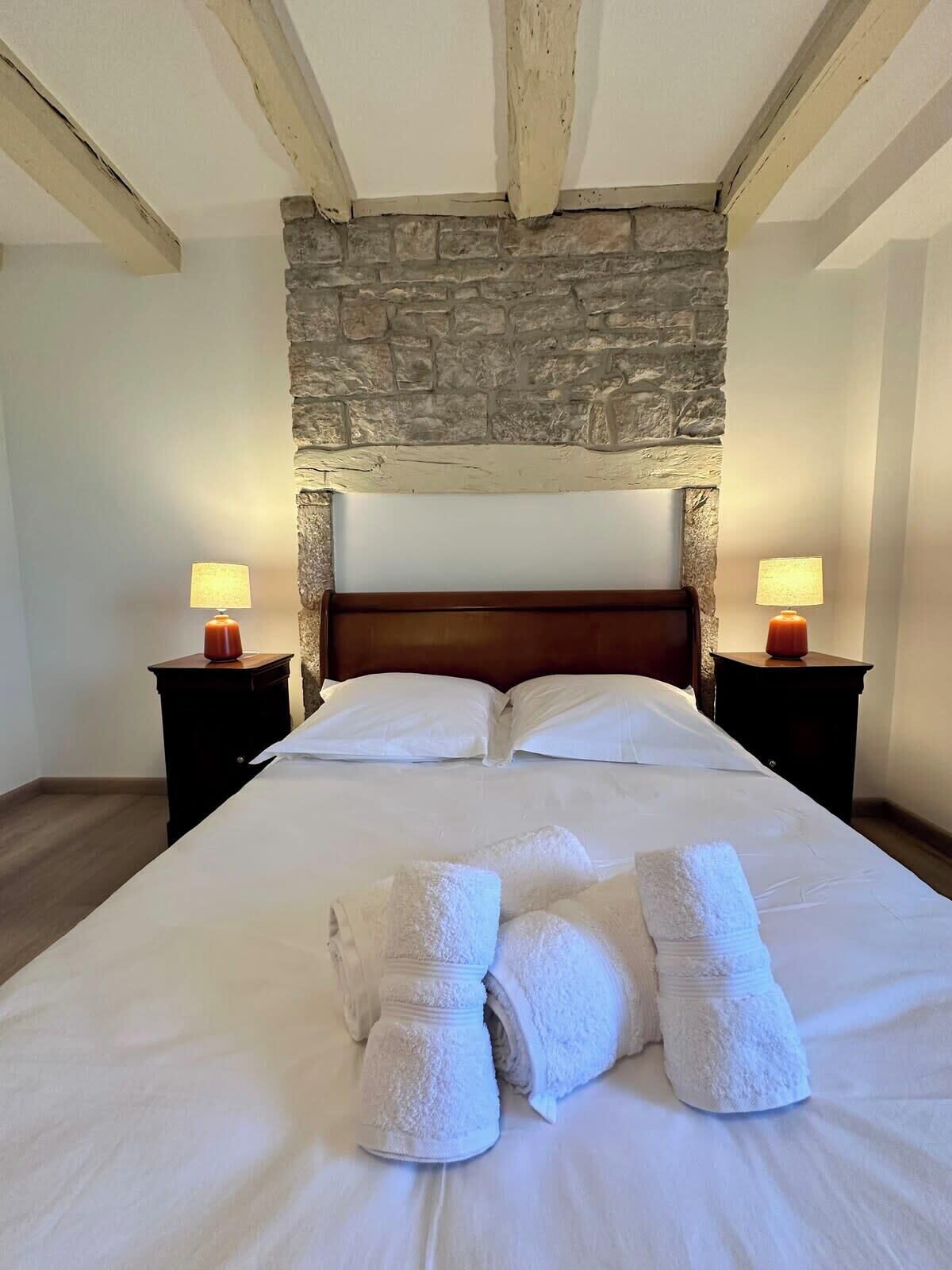 Cama bem arrumada com lençóis e toalhas brancas, parede de pedra acima e duas luminárias de cabeceira em mesas de criado-mudo escuras.