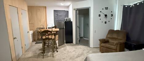 A room with a bed, a chair, a dining table with chairs, a refrigerator, a clock on the wall, and a door leading to another room.