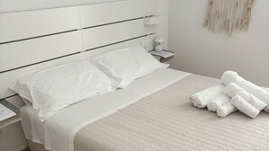 Een keurig opgemaakt bed met witte lakens en een beige gebogen deken, een nachtkastje met een lamp en een decoratie aan de muur.