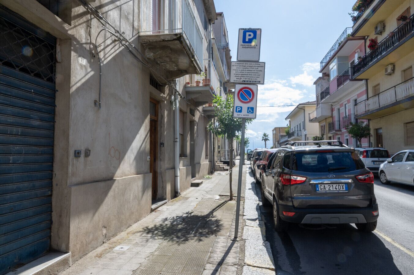 Une rue étroite bordée de voitures garées, une porte de garage bleue et un bâtiment sur lequel est apposée une enseigne indiquant un espace de stationnement.