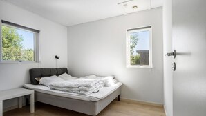 Un dormitorio minimalista con una cama individual, un banco y dos ventanas que dejan entrar luz natural.