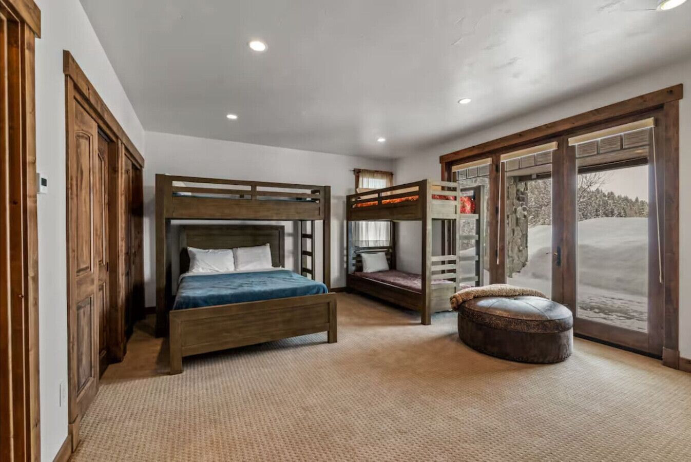 A bedroom with a bunk bed, a single bed, a large window with a view of a snowy landscape, and a wooden door.