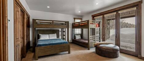 A bedroom with a bunk bed, a single bed, a large window with a view of a snowy landscape, and a wooden door.
