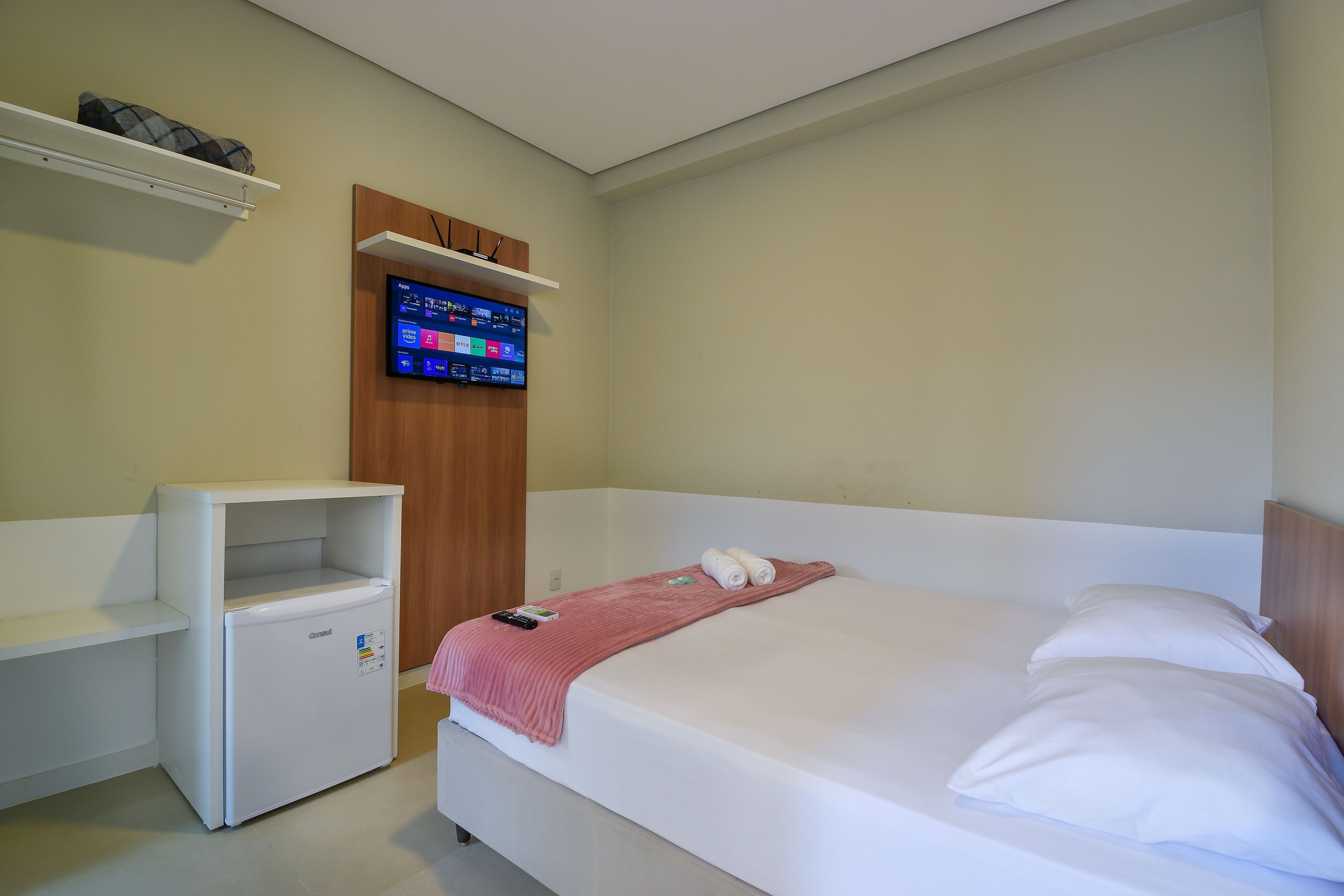 A bedroom with a bed, a small white desk, a television mounted on the wall, and a wooden wardrobe.