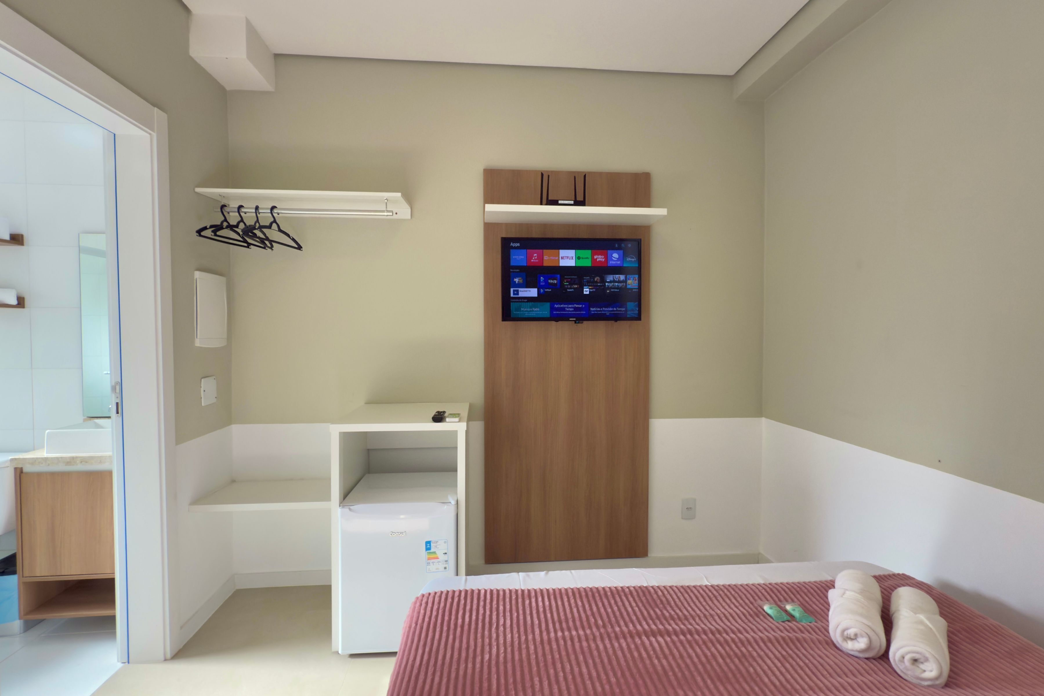 Una habitación de hotel moderna con un armario empotrado, un televisor de pantalla plana colocado encima y una cama con colcha rosa, junto con una mesita de noche pequeña con un cepillo de dientes y pasta dental.