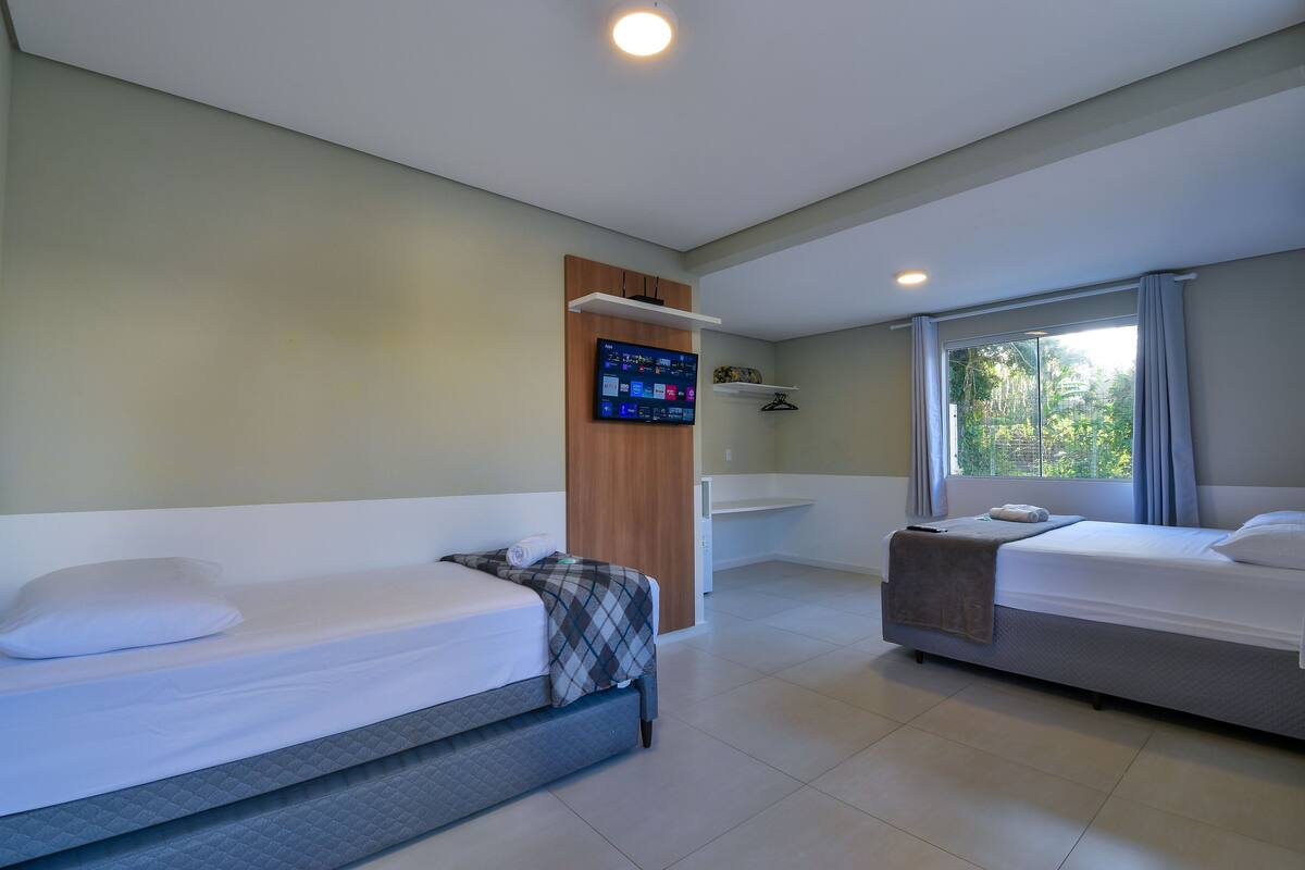 A hotel room with two beds, a TV mounted on the wall, a window with curtains, and a small shelf with a towel.